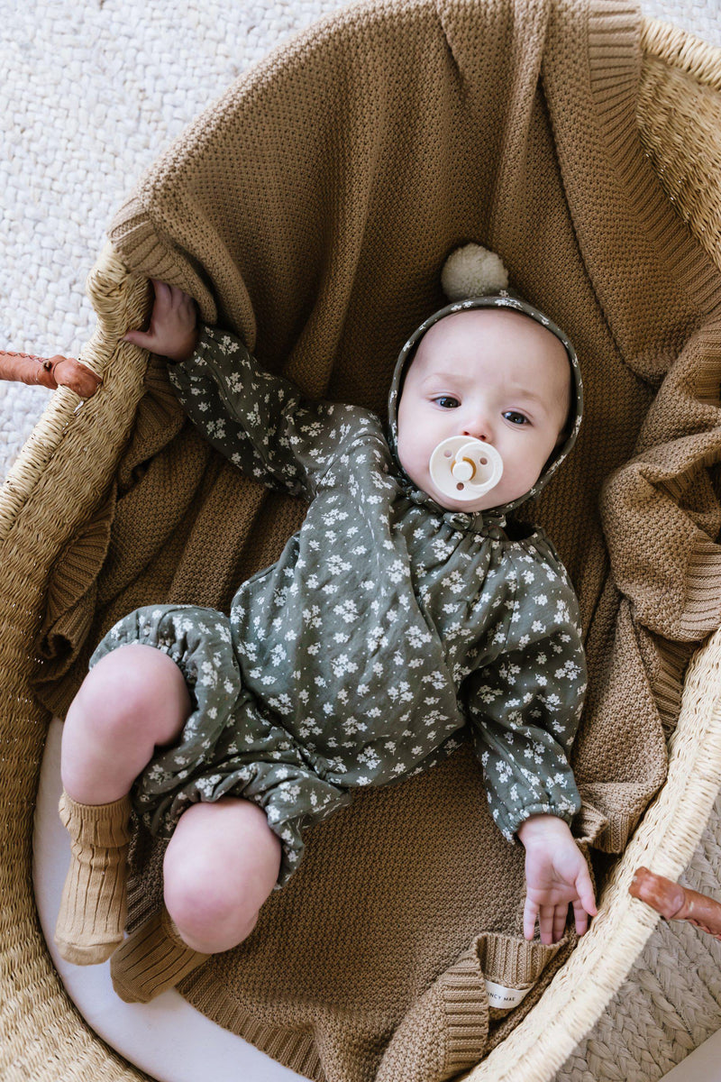 Quincy Mae Fall 2020 collection pieces - baby on clay chunky organic knit baby blanket wearing woven cinch longsleeve tee and bloomers with knee socks and pom pom bonnet