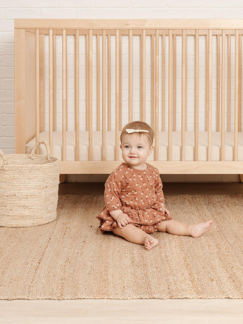 Little girl sitting in front of a wooden crib wearing the Quincy Mae Rosie Romper in Clay Ditsy, with a little knot headband in Natural. 