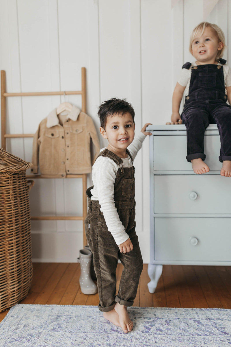 Two boys wearing the Jamie Kay Marigold collection Jordie corduroy overalls in cedar and Peacock; one boy sitting on a dresser and the other boy standing on the rug. The Will Cord Jacket in almond is behind them hanging on a wooden drying rack.