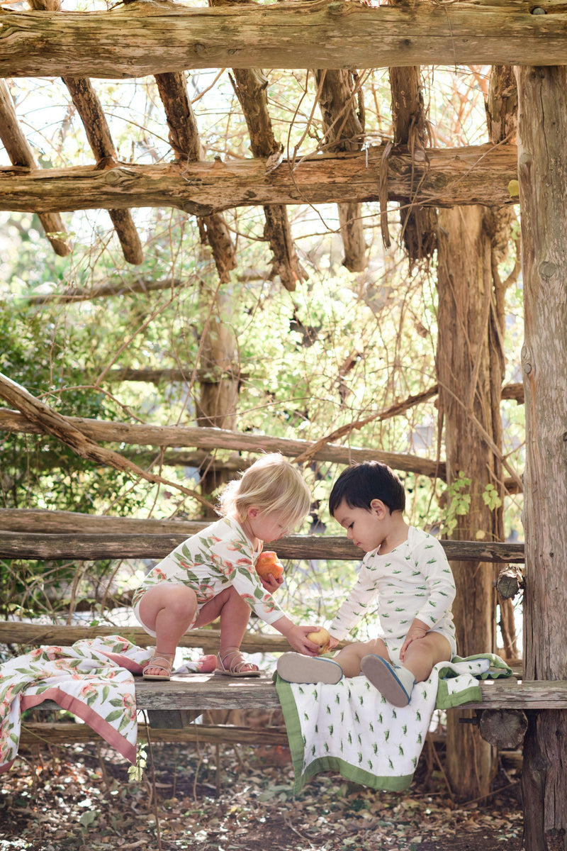 MilKBarn Kids lifestyle shot of two toddlers with a peach wearing new organic cotton grasshopper and peaches print long sleeve bodysuits next to the Big Lovey Blanket in grasshopper and peaches outside under a wood canopy. 