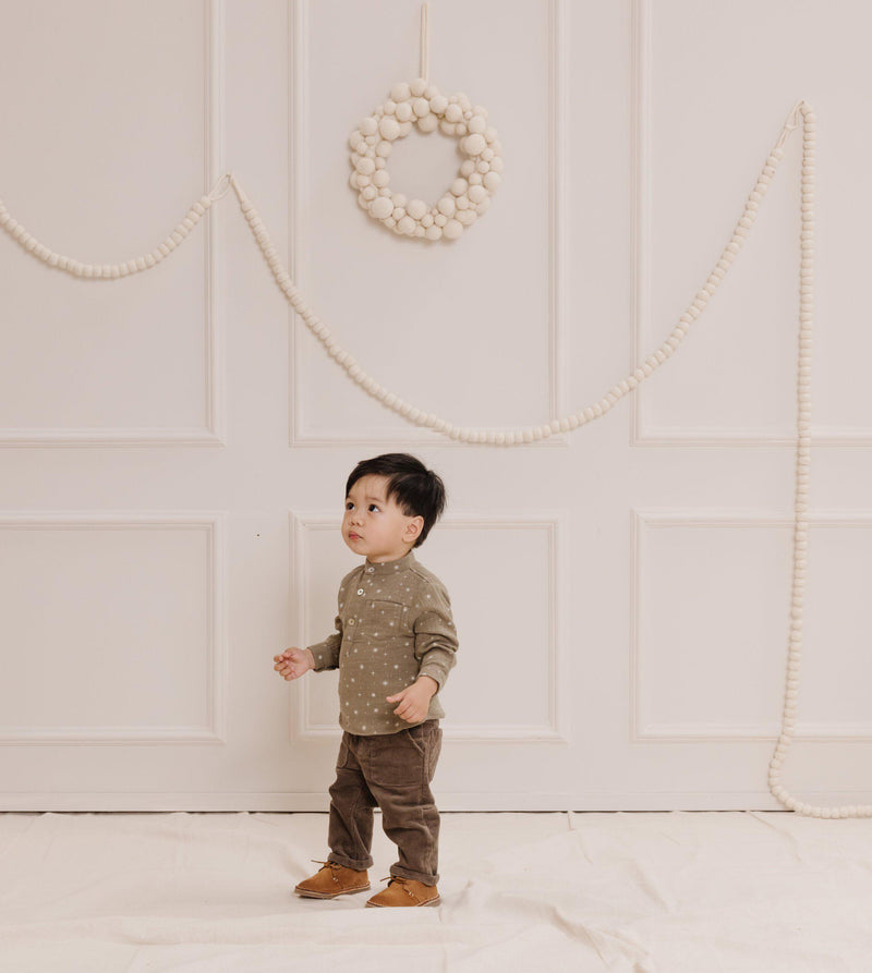 Little boy wearing the Rylee + Cru Holiday Collection Mason Shirt in Stars with Zander pants in Olive. Lifestyle photoshoot standing in a room with wood paneling, a white Christmas wreath, and some white garland draped across the wall. 