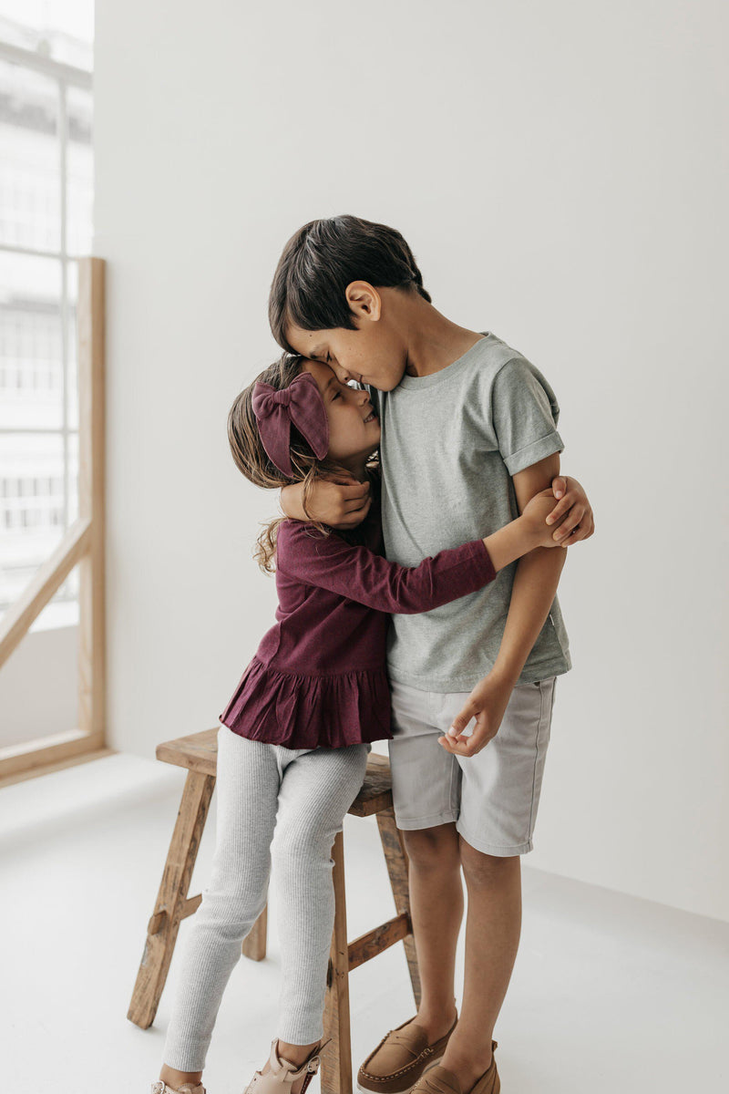 Lifestyle photo for Jamie Kay clothing - little girl wearing Plum Bailey long sleeve top and oatmeal marle essential leggings hugging brother wearing Norway Sam tee while sitting on wooden stool in studio. 
