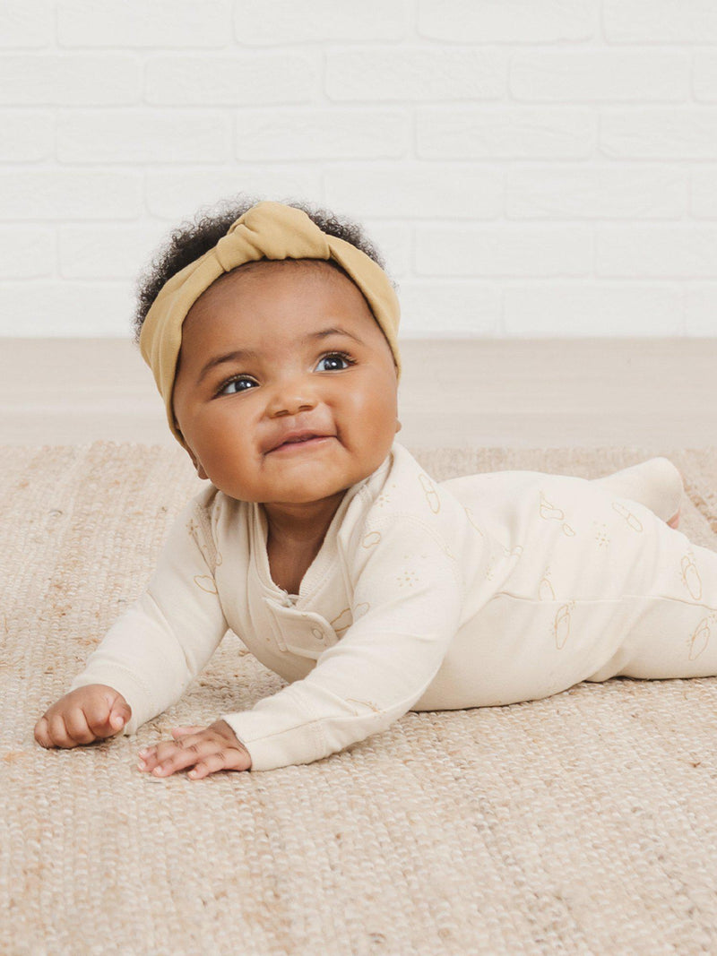 Quincy Mae Spring Summer 2021 Drop 1 arrives Friday, May 14. Photo is of a baby girl laying on her stomach smiling up at the camera wearing the gold headband and the new zipper sleeper in the clouds print. 